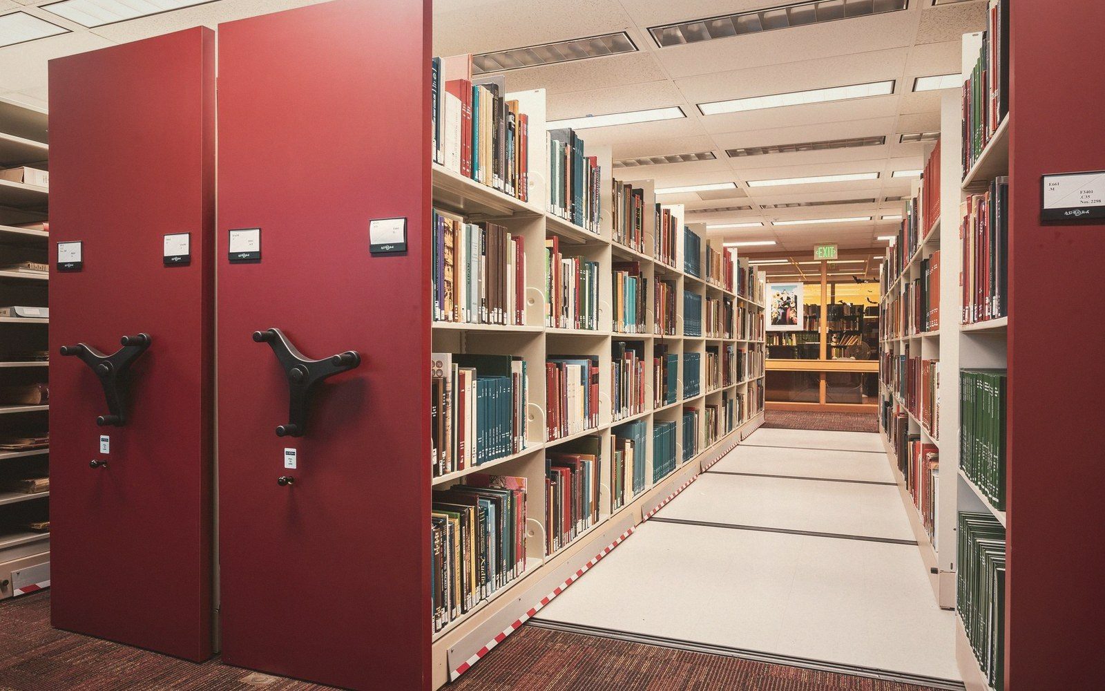Rows of books on movable compact shelving units with red end panels and hand cranks