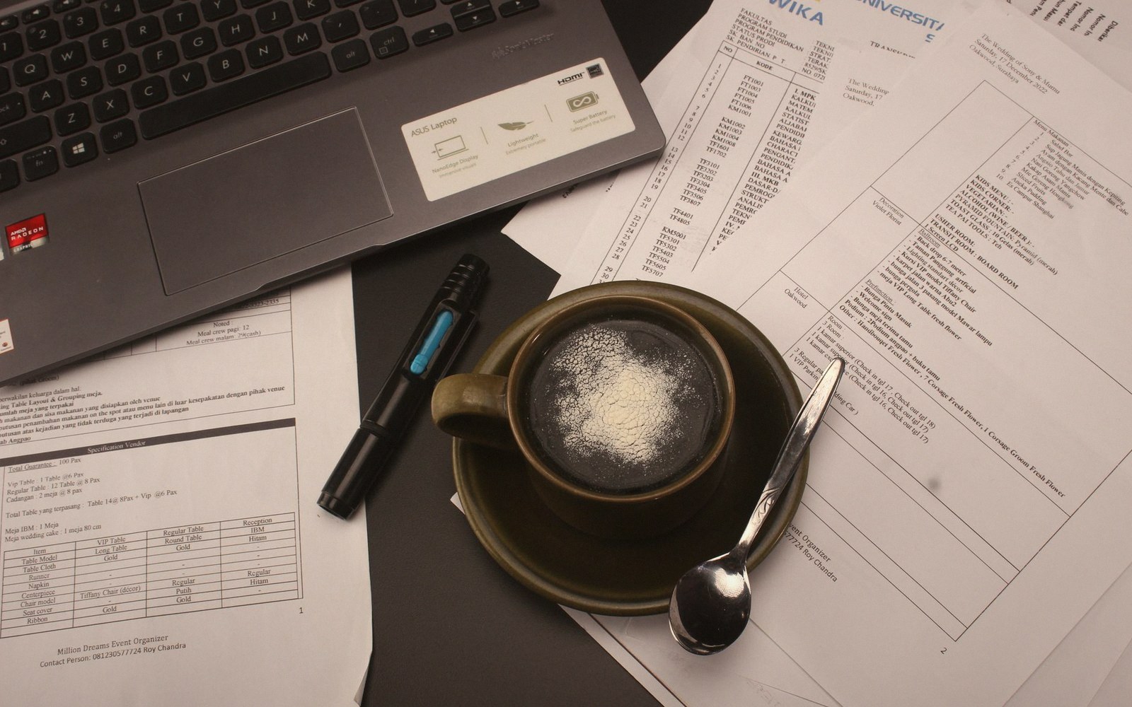 A coffee cup, spoon, pen and laptop surrounded by printed spreadsheets on a desk